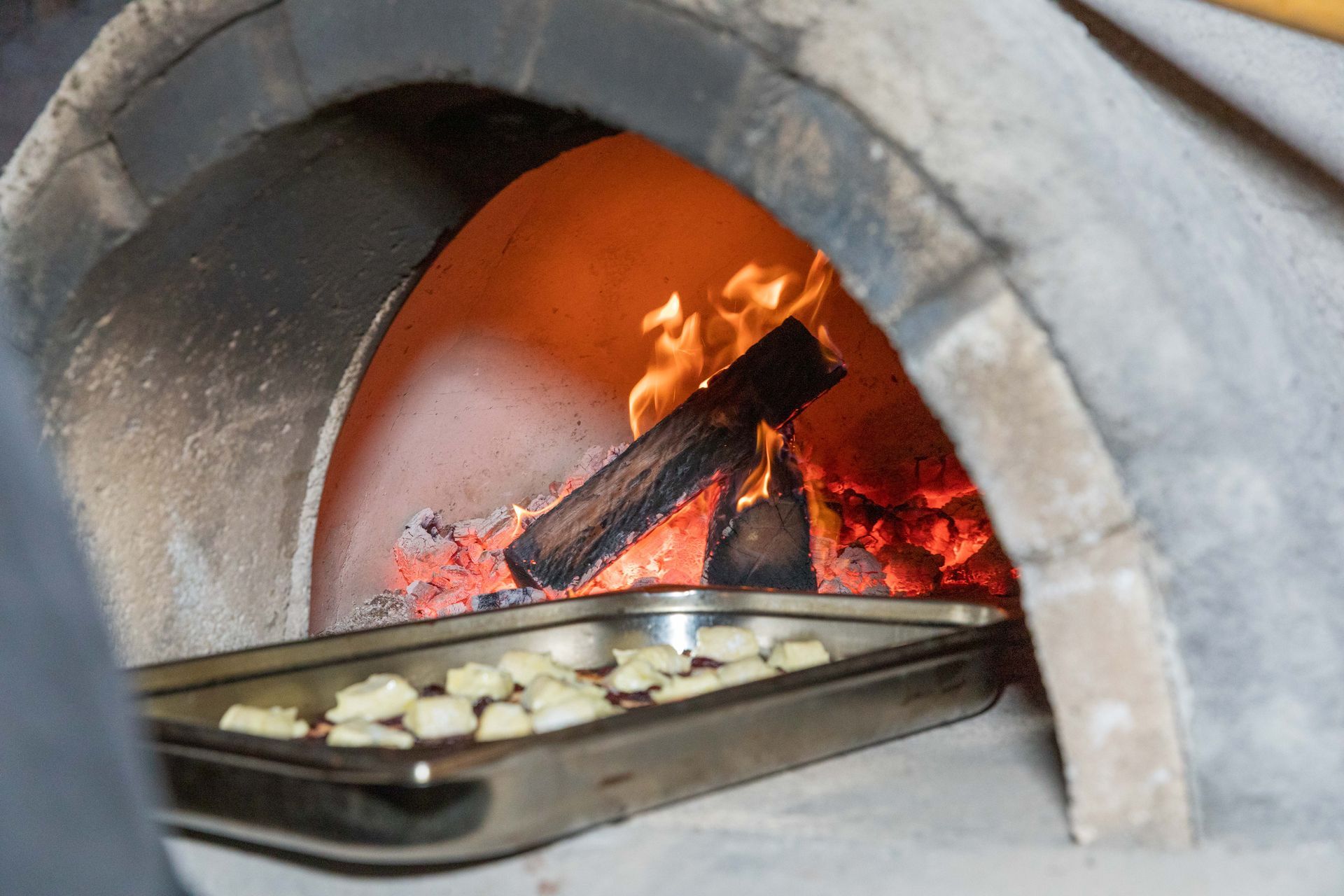 A tray of food is placed in a wood-fired oven, with flames and burning wood visible.