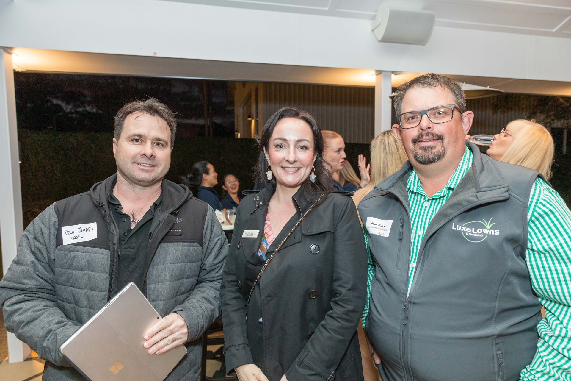 Three people smiling at an outdoor gathering. A man on the left holds a laptop, the woman in the centre wears a black jacket, and the man on the right wears a vest.