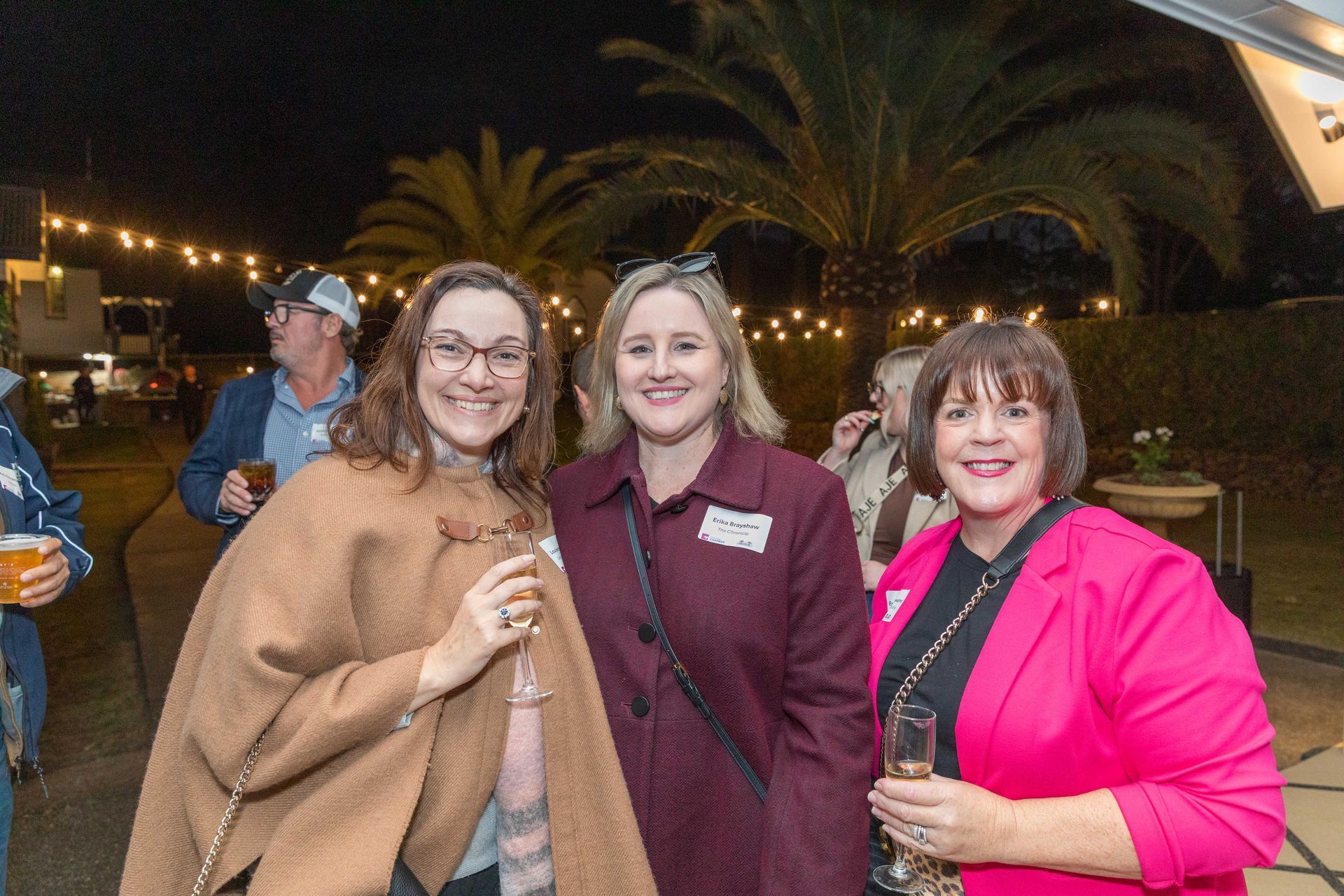 Three women smiling outdoors at night, one wearing a pink blazer, another a burgundy coat, and the third, a brown shawl.