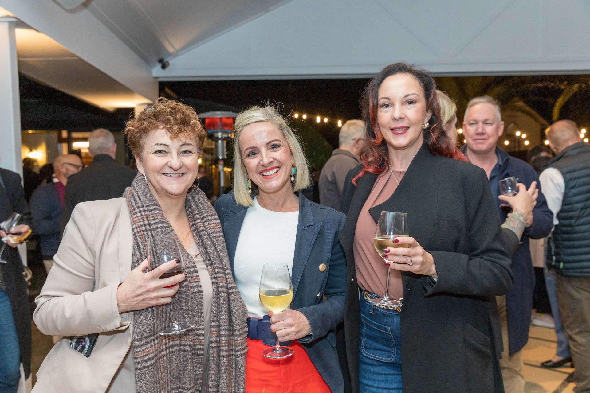 Three smiling women holding drinks at an outdoor evening event. Others in the background.