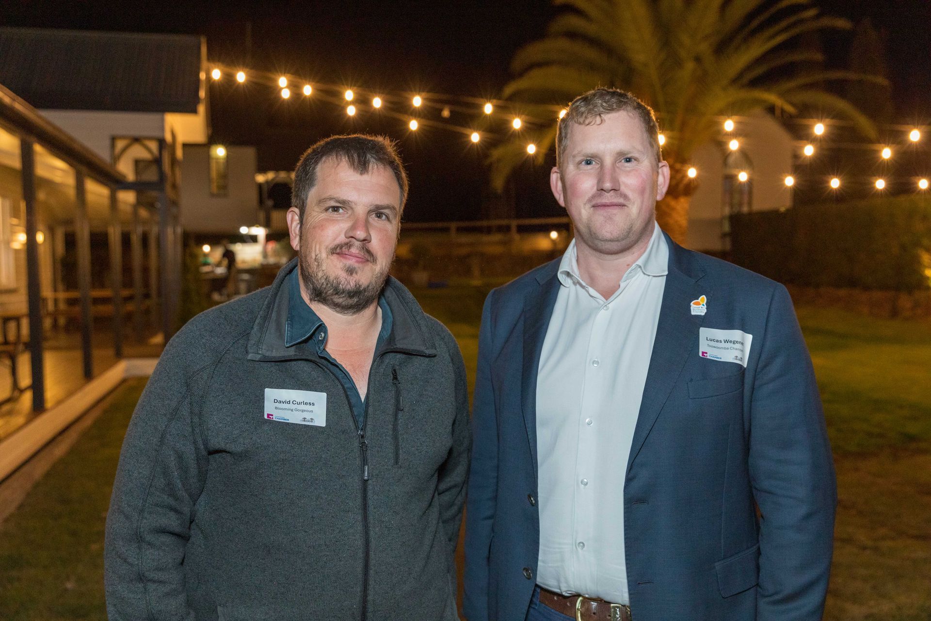 Two men standing outdoors at night, smiling, with string lights and a building in the background. One wears a jacket, the other a suit.