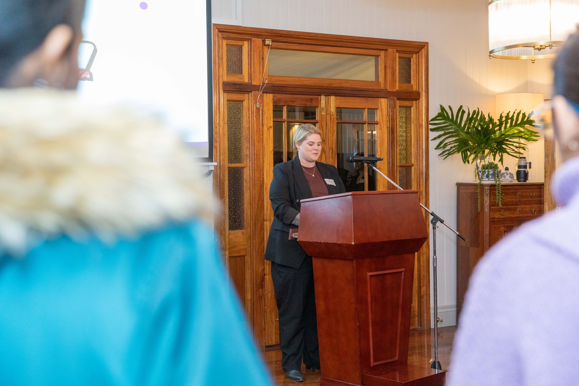 A woman in a suit speaks at a podium in front of a wooden door, addressing an audience.