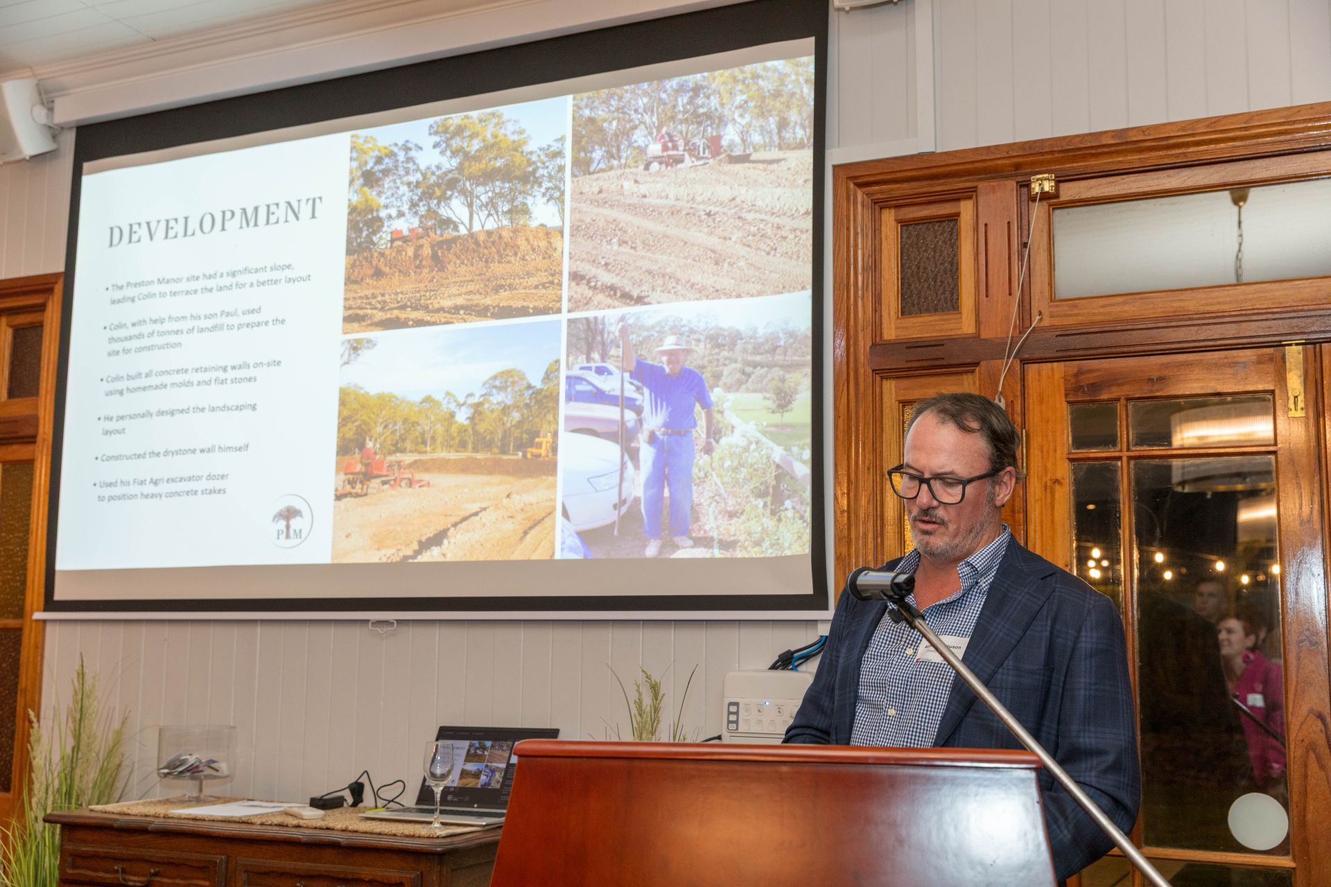 A man in a blazer speaks at a podium; a presentation screen behind him shows development images. Interior setting with wooden accents.