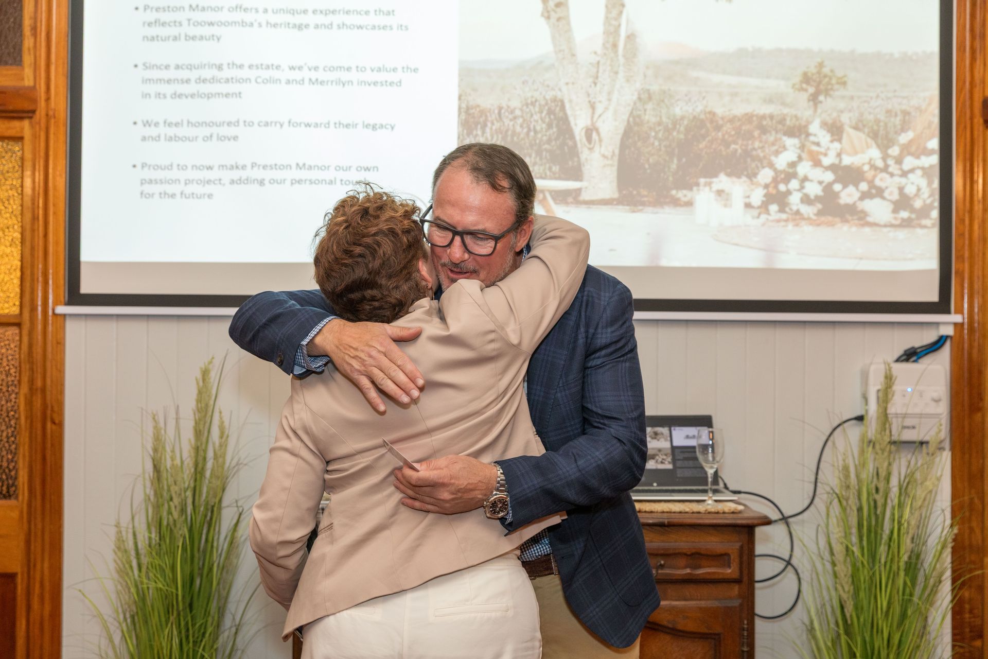 Man in blue blazer hugging woman in tan jacket; a presentation screen and floral decorations are in the background.