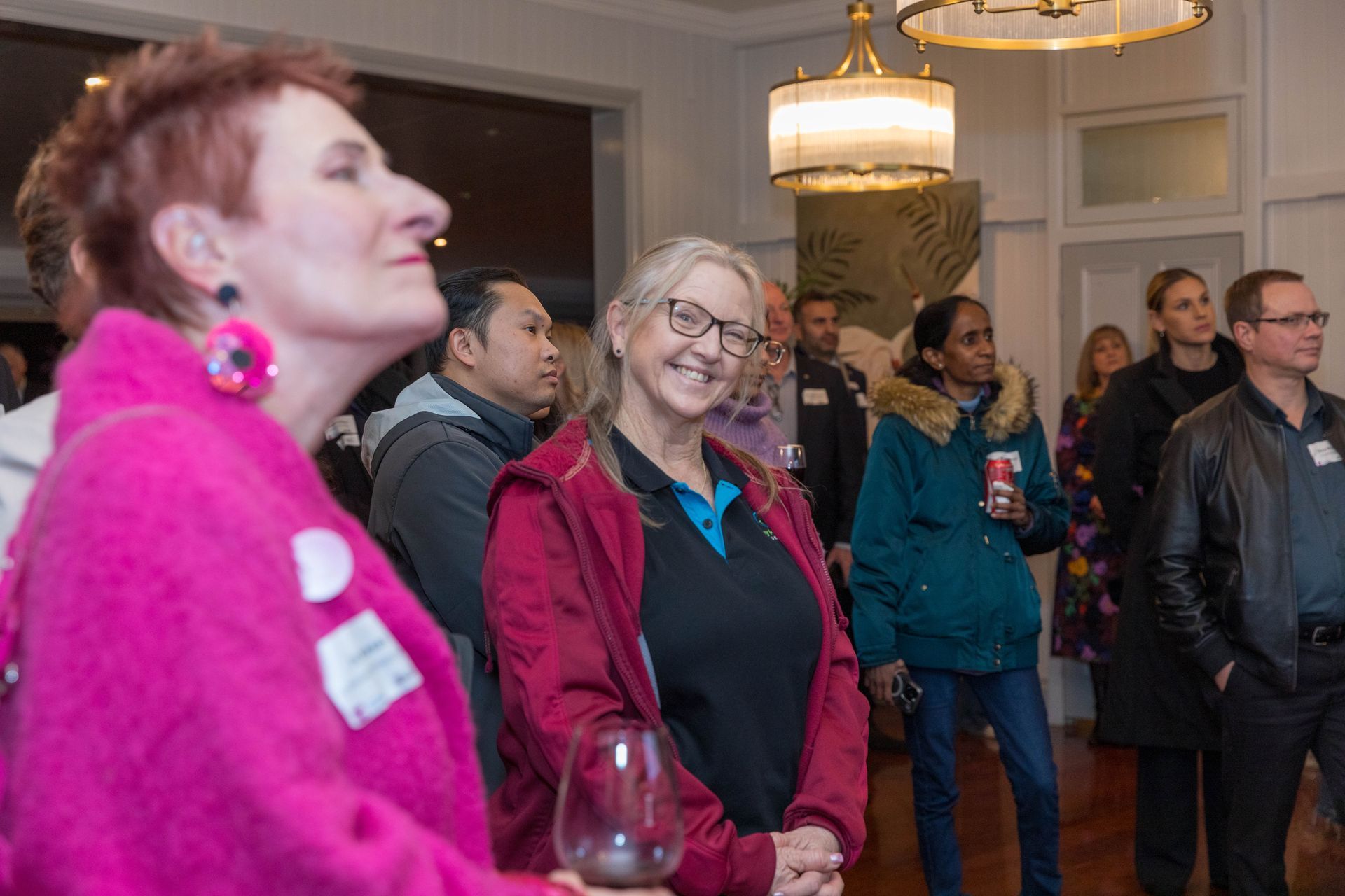 Attendees at a social event; woman in pink jacket in foreground, others looking on, some smiling. Well-lit interior with chandelier.