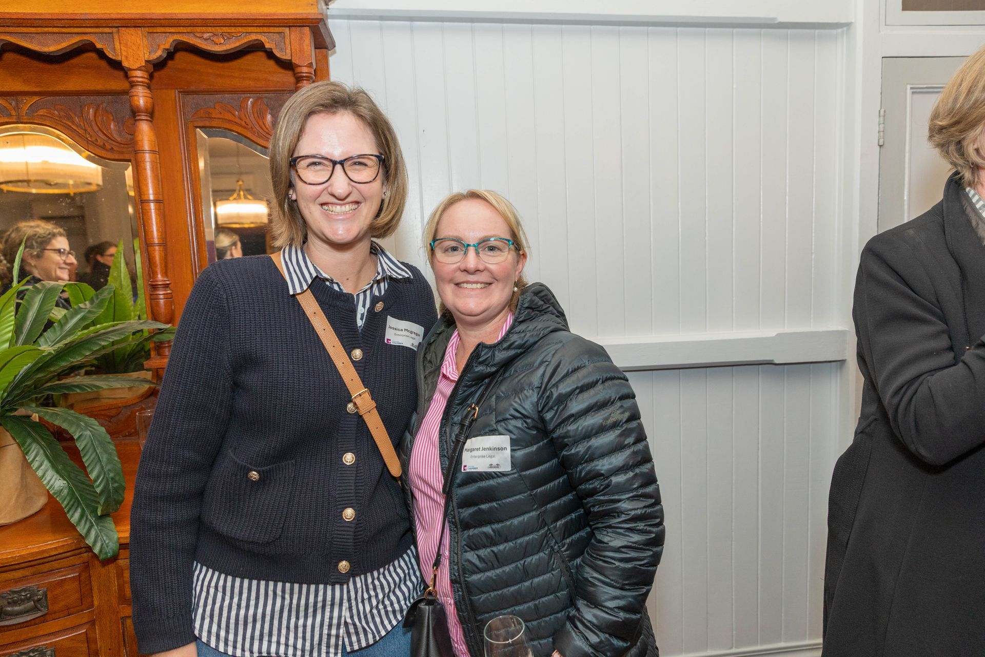 Two women smiling, posing together indoors. One wears glasses and a navy sweater, the other a pink shirt and puffer jacket.