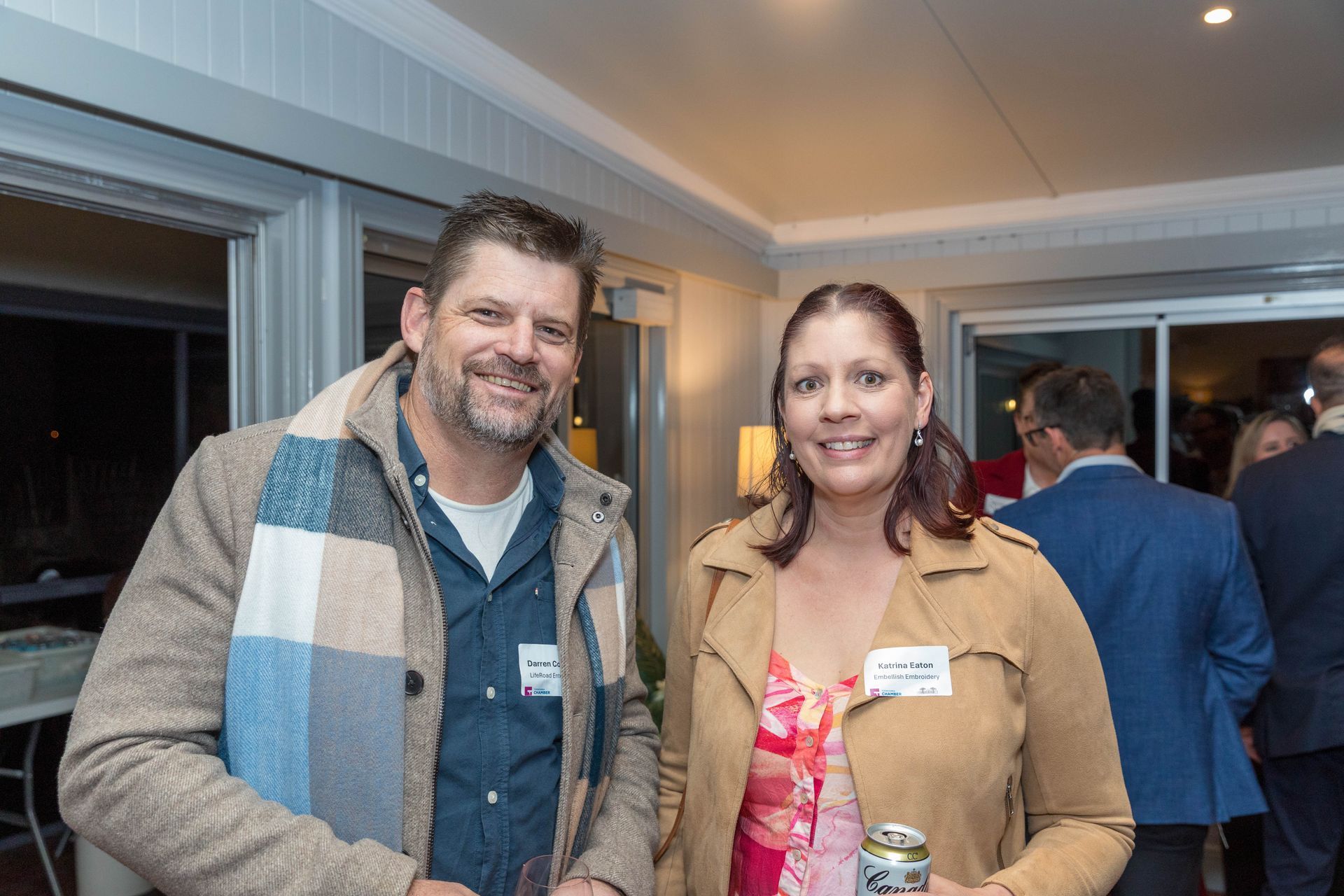 Man and woman smiling at a social event. Man wears a scarf and jacket, woman a light-brown jacket and floral top.