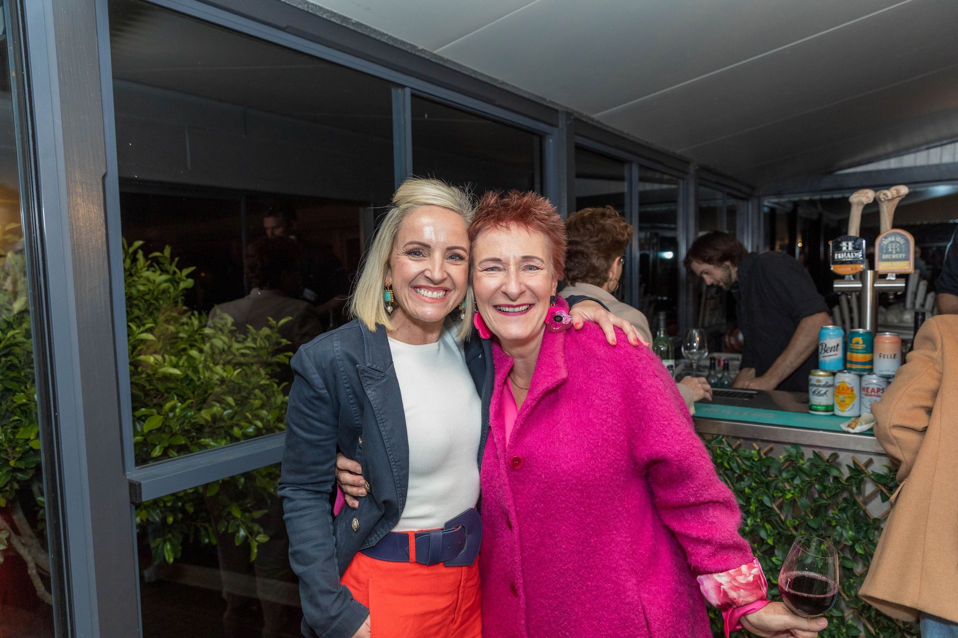 Two smiling women pose together at an outdoor event. The woman on the left wears a blue jacket, white shirt, and orange shorts. The woman on the right wears a pink coat.