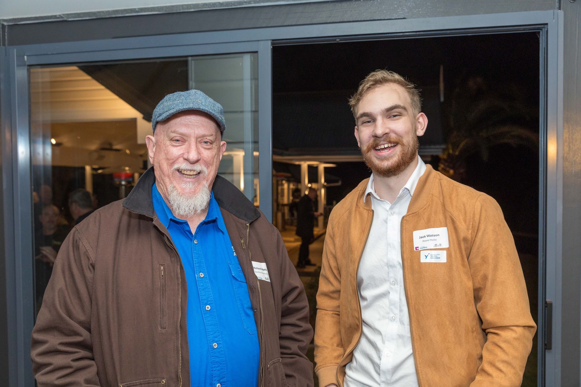 Two men smiling near a sliding glass door. One man wears a brown jacket, blue shirt, and a cap. The other wears a tan jacket and white shirt.
