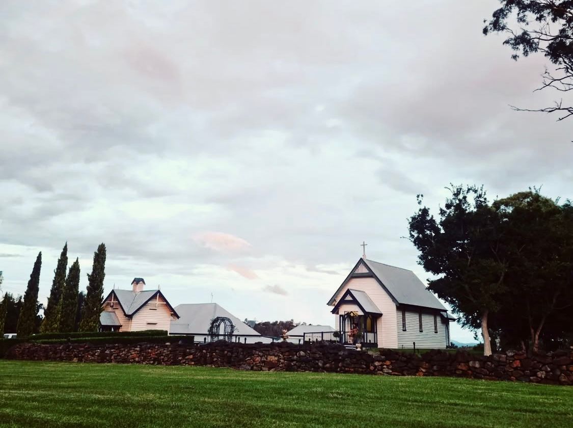 A small chapel and building with a stone wall are set against a cloudy sky, on a green lawn.