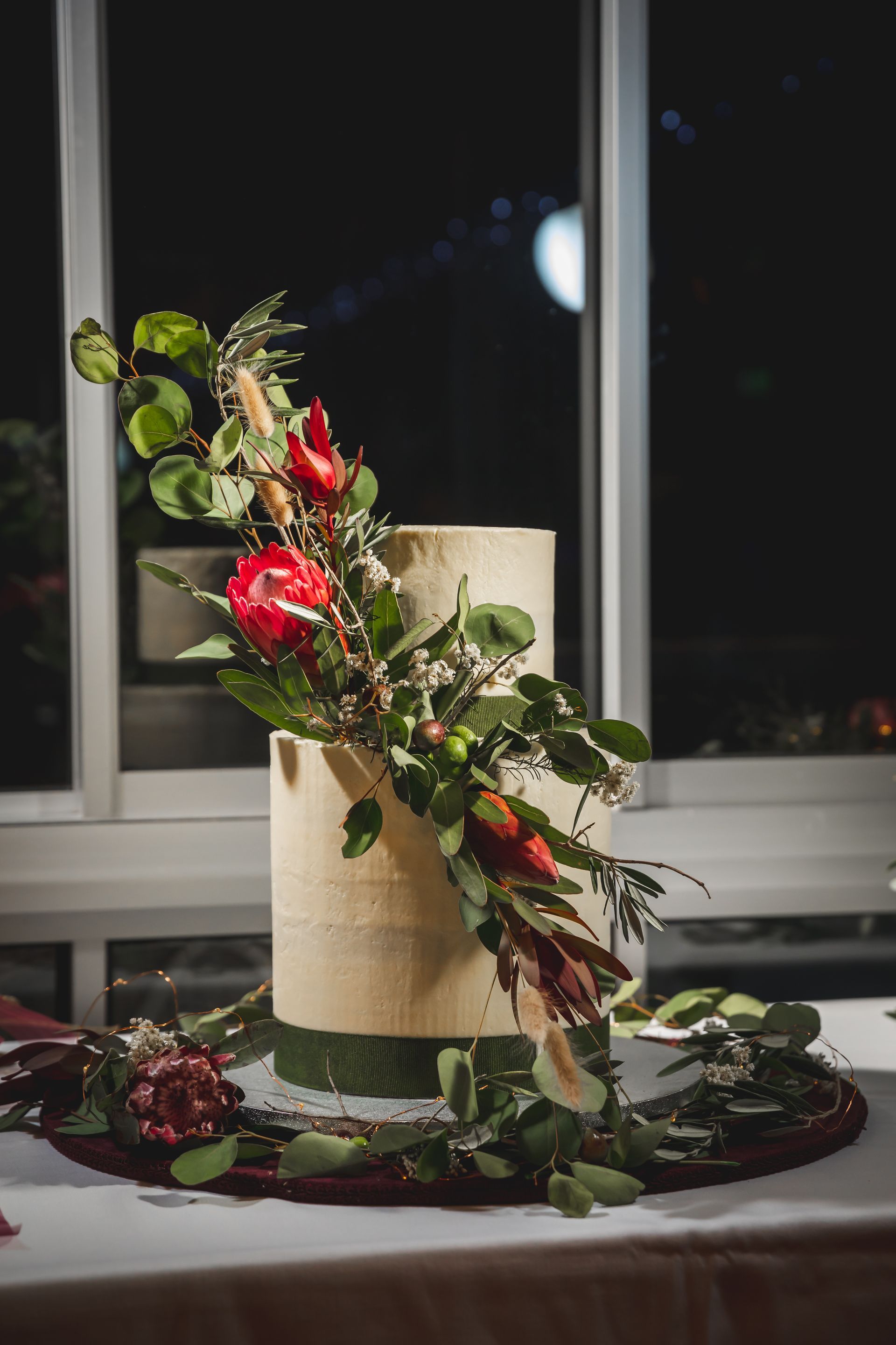 Two-tiered cake decorated with greenery, red flowers, and burlap texture, on a table near a window at night.