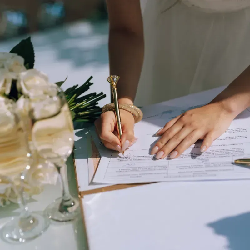 A bride signs a document with a gold pen at a table with champagne glasses and a bouquet of white flowers.