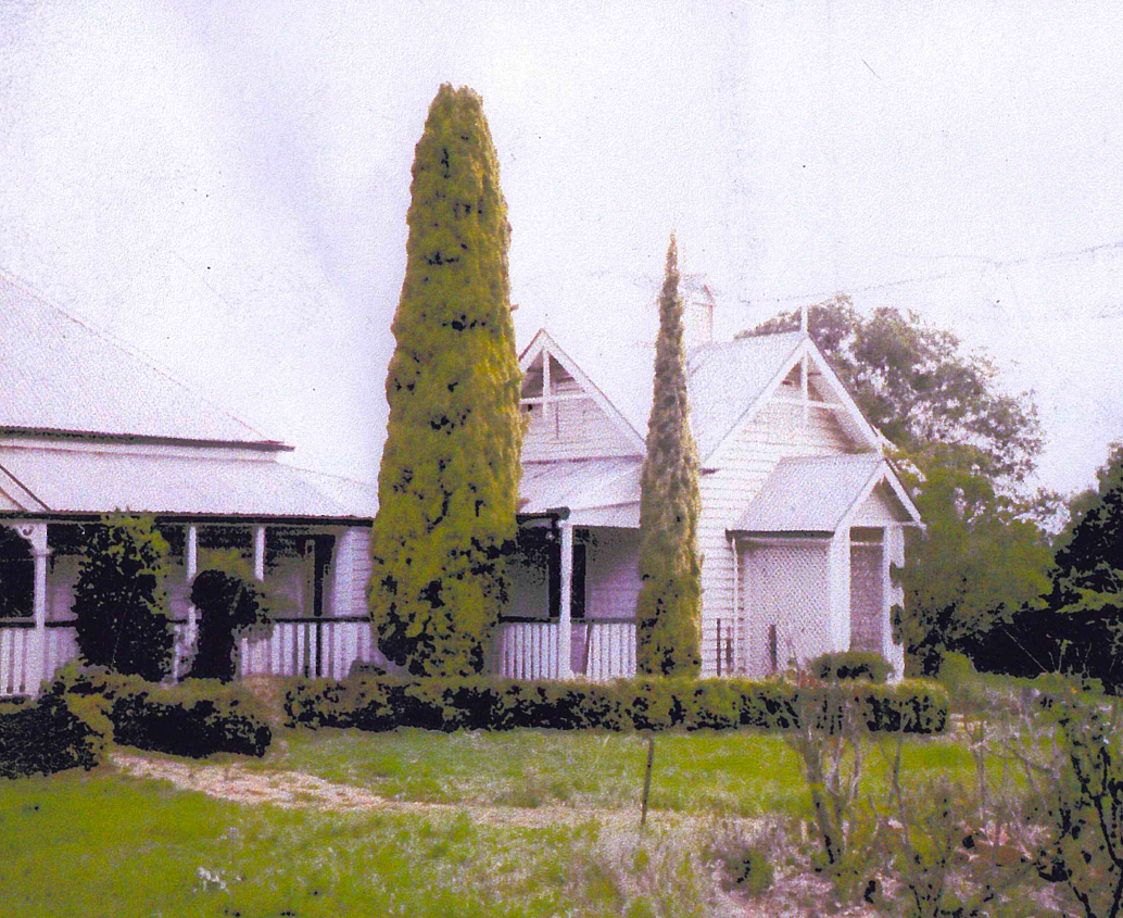 White house with a porch, two tall trees, and a green lawn. The house has a gabled roof and appears to be in a rural setting.