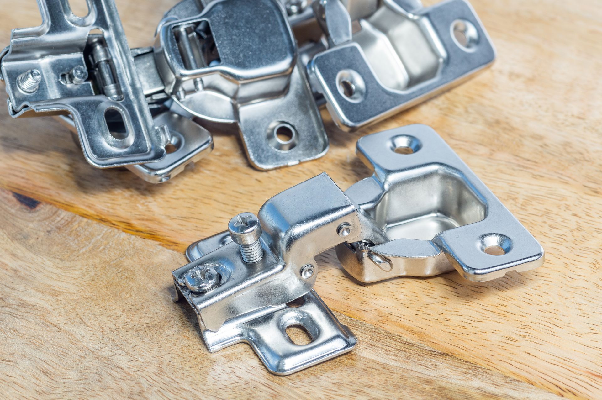 Close-up of metal cabinet hinges and mounting plates arranged on a wooden surface. Close-up of metal cabinet hinges and mounting plates arranged on a wooden surface.