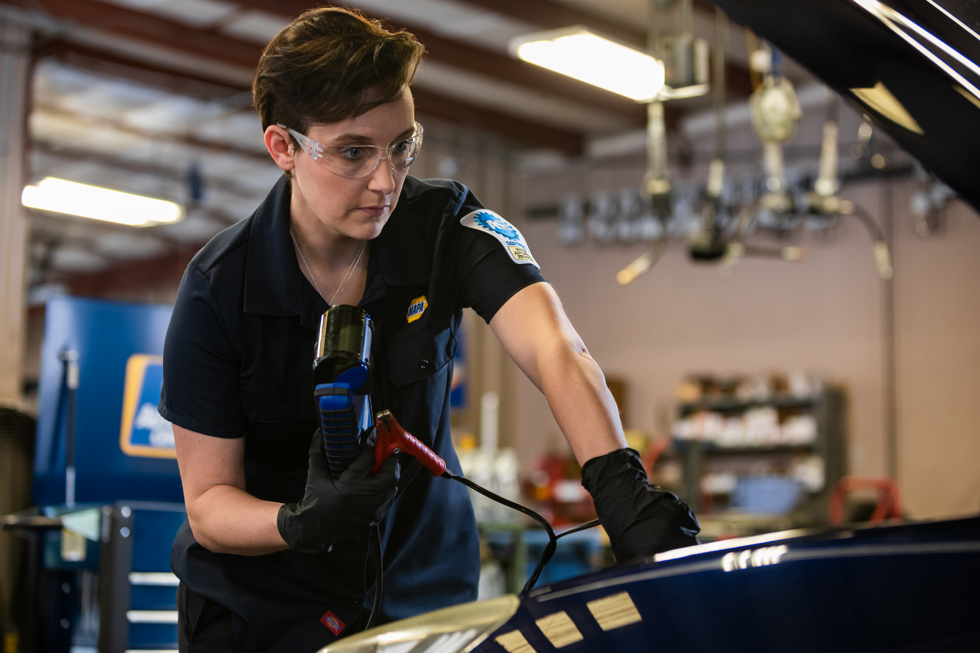 Technician in safety glasses and gloves inspects a car in a workshop with a diagnostic tool.