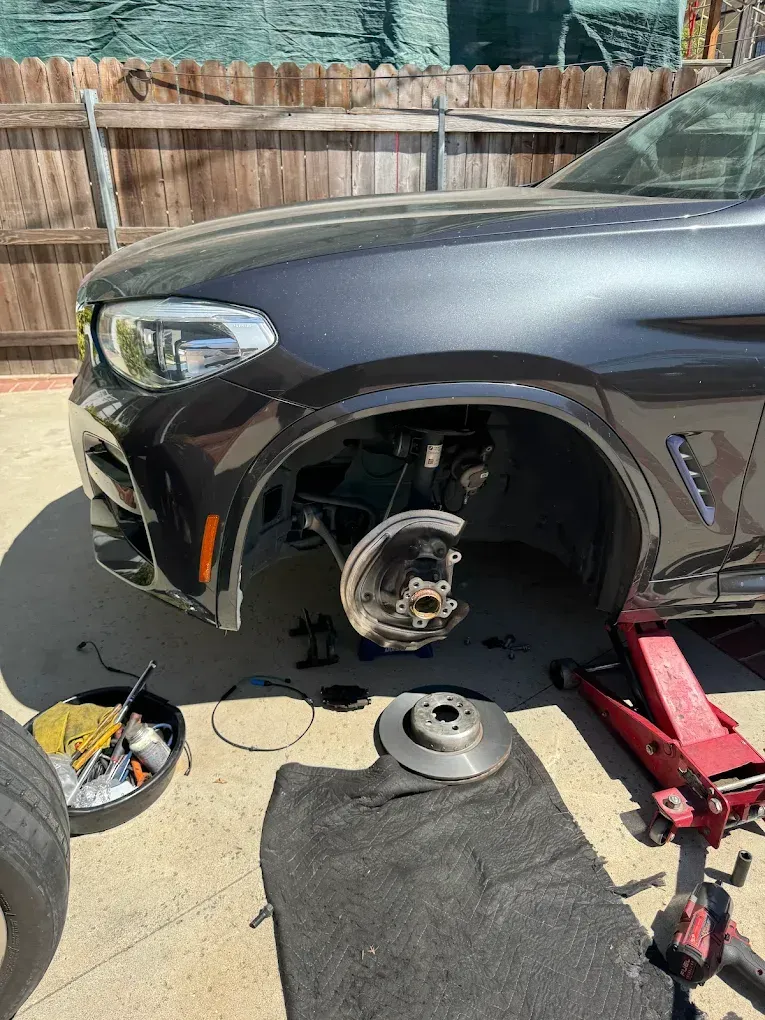 A dark gray BMW with its front wheel removed, brake rotor on the ground, jack beneath.