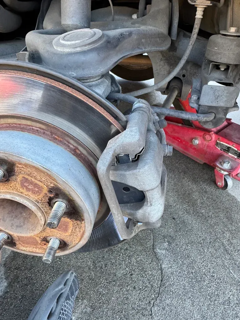 Car wheel with brake rotor, caliper, and suspension components, jack in background.