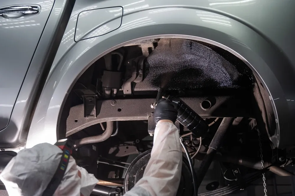 Person spraying undercarriage of a silver vehicle with black coating, likely for rust protection, in a workshop setting. — Pelican Motors Service Centre in Caloundra West, QLD