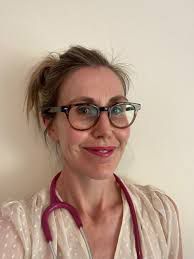 Person wearing glasses and a pink stethoscope, smiling against a plain wall.