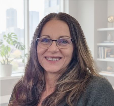 Smiling woman with glasses in a bright room, seated near a window with plants in the background