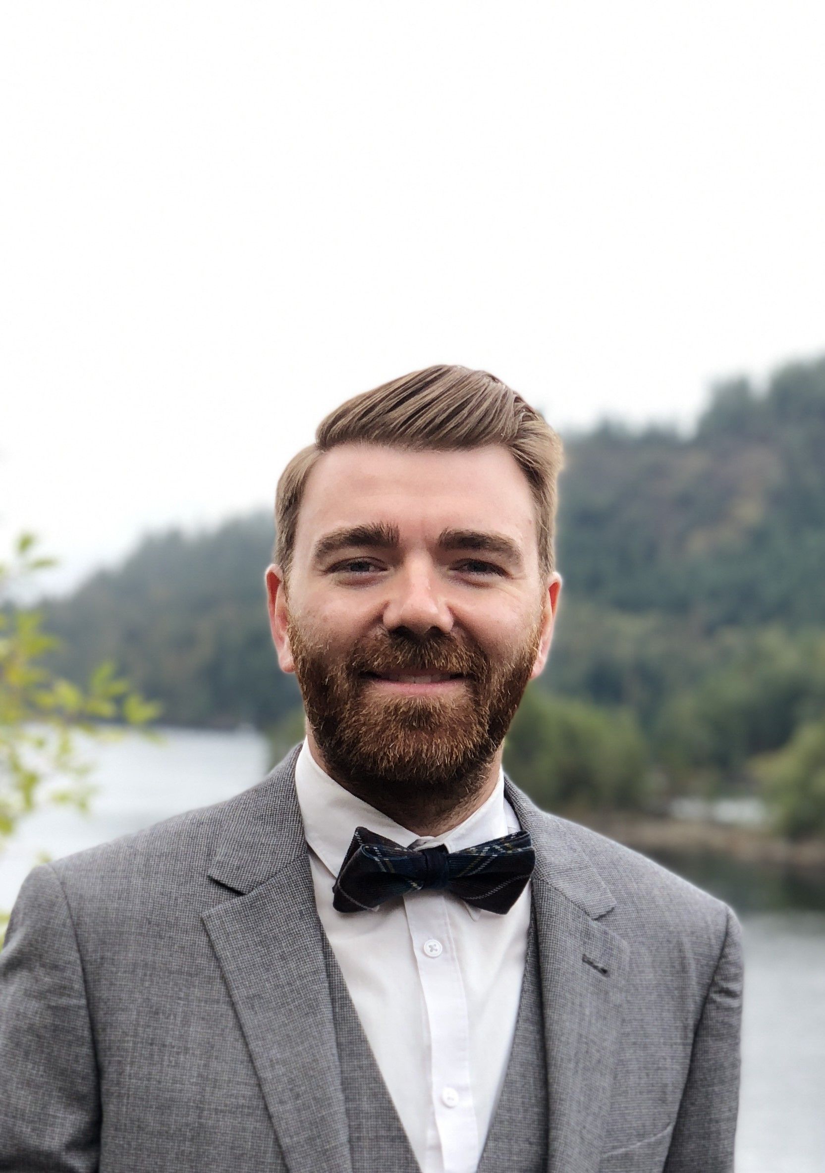 Man in a gray suit and black bow tie smiling outdoors with a blurred lake and trees behind him
