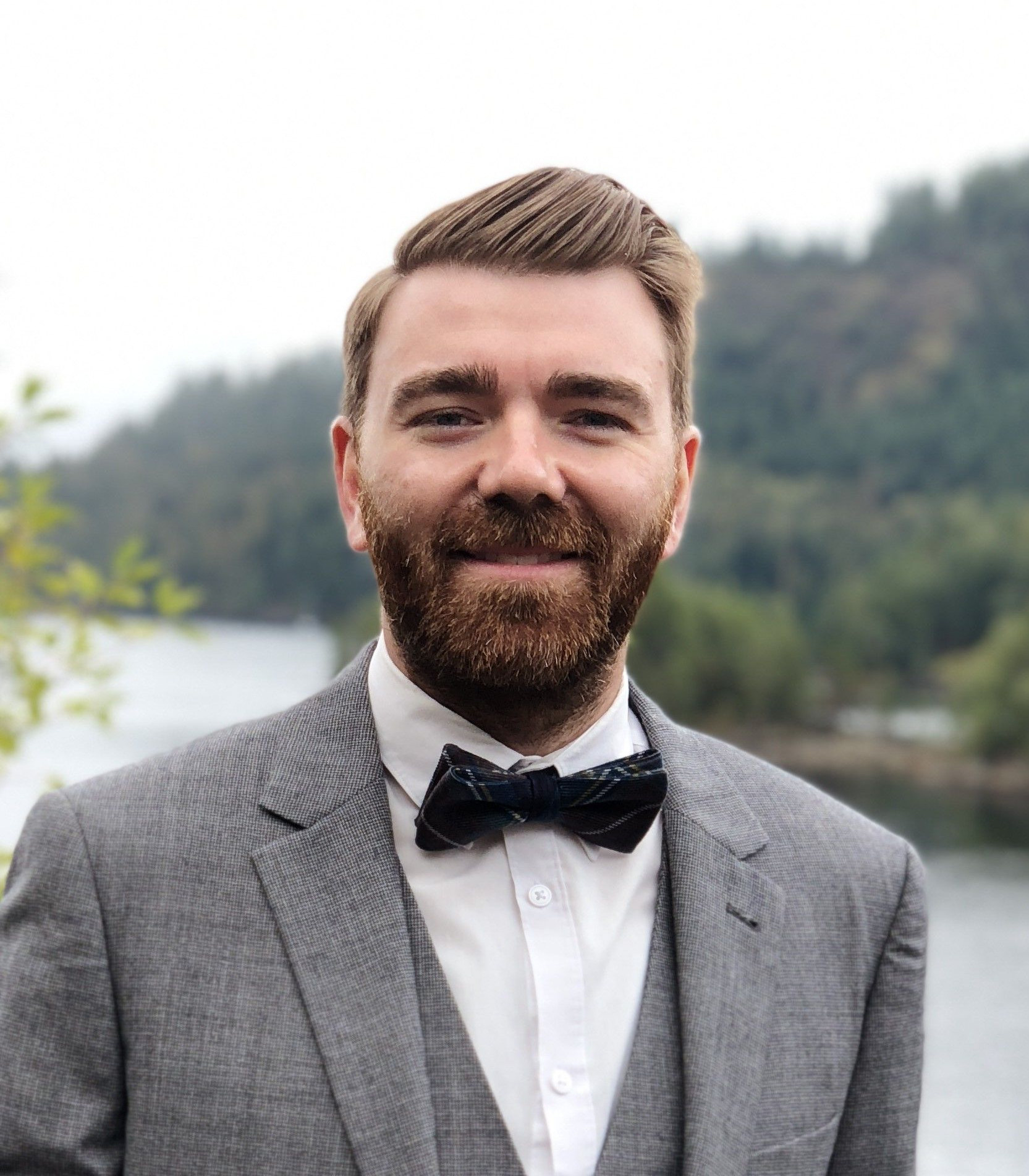 Smiling bearded man in a gray suit and bow tie outdoors with trees and water behind him