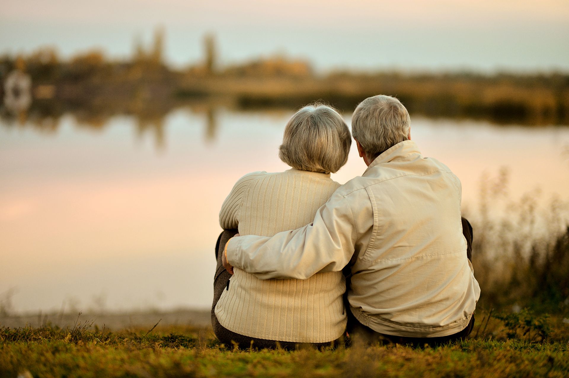 Two people sit close together on a grassy shore, facing a calm lake at sunset.