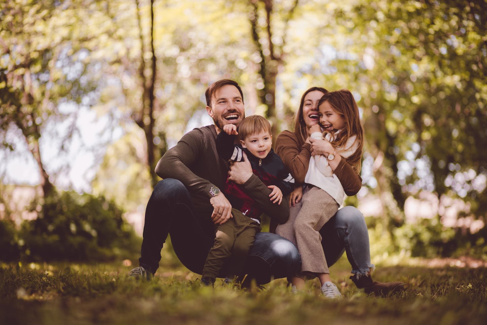 Family crouching in a sunlit park, smiling and hugging among autumn trees.
