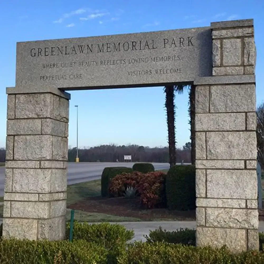 Stone entrance arch for Greenlawn Memorial Park with landscaped shrubs and road in the background