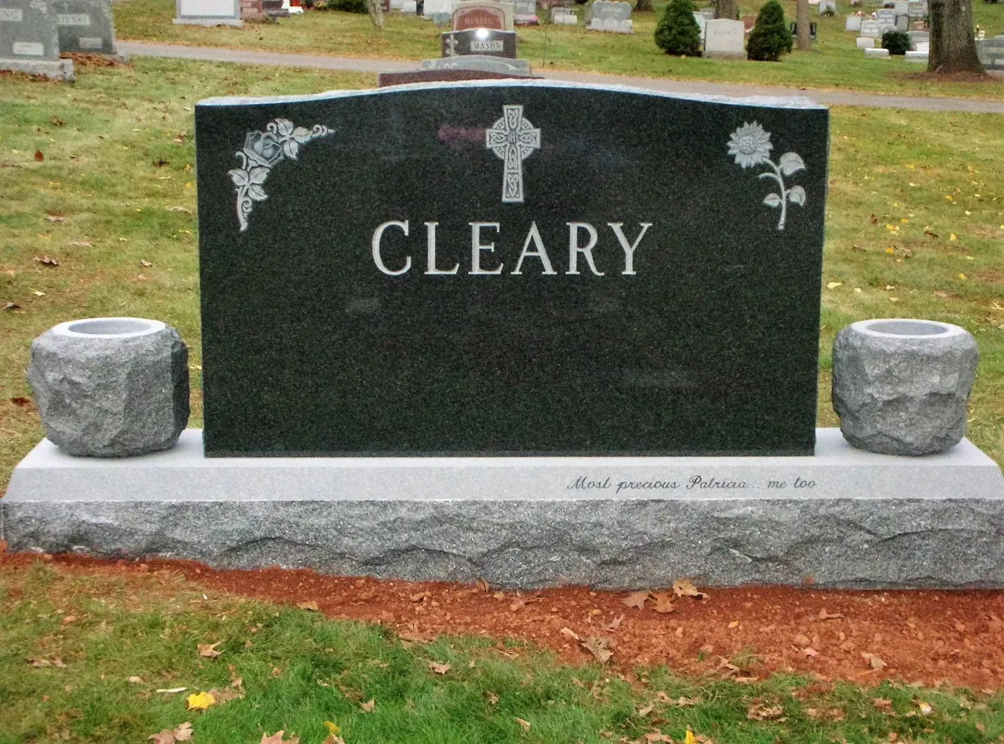 Black granite gravestone engraved “CLEARY” with flower carvings and two small stone vases on a cemetery plot