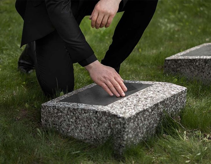 Person kneels by a granite gravestone, placing a hand on the plaque in a grassy cemetery