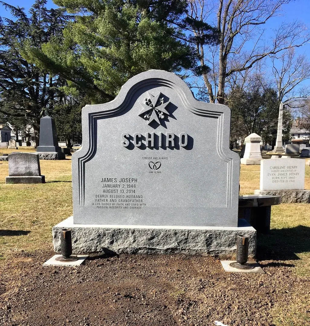 Gray memorial headstone in a cemetery, with engraved emblem and name, against trees and blue sky