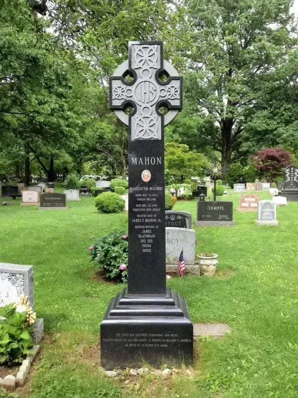 Tall black Celtic cross gravestone in a green cemetery with other headstones and trees in the background