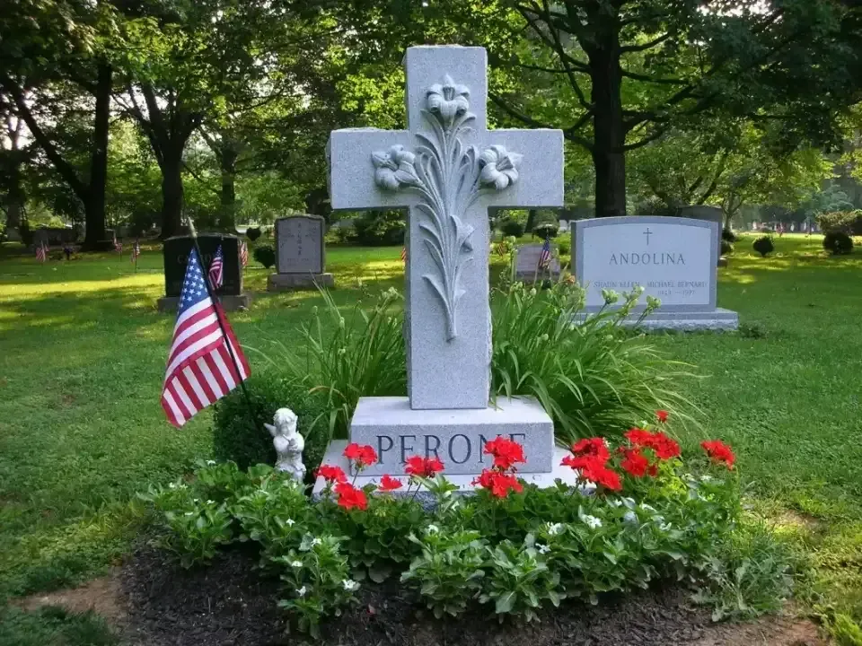 White cross gravestone with red flowers and a small American flag in a green cemetery.