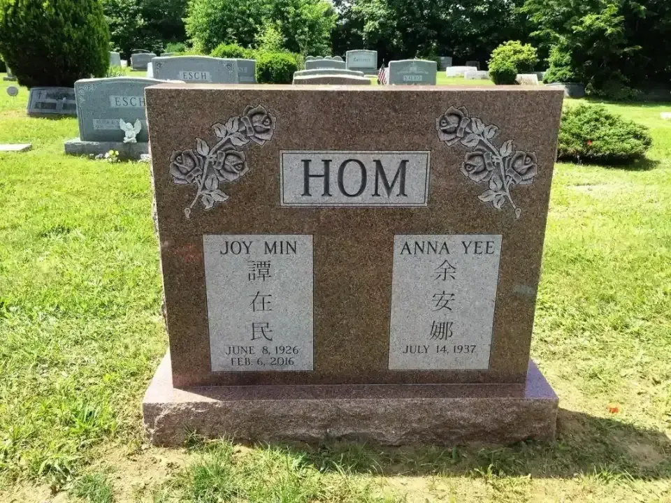 Brown gravestone monument with “HOM” and engraved Chinese text in a grassy cemetery.