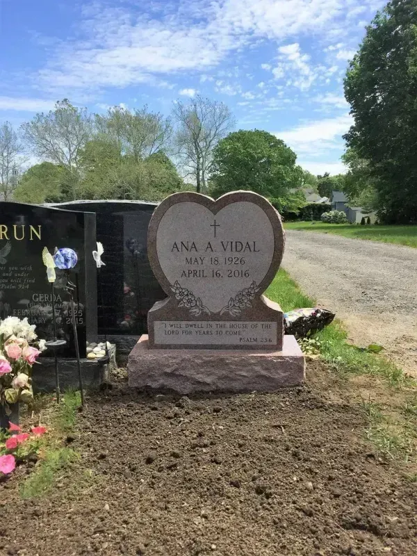 Heart-shaped gravestone in a cemetery beside a road, with flowers and fresh soil under a blue sky.