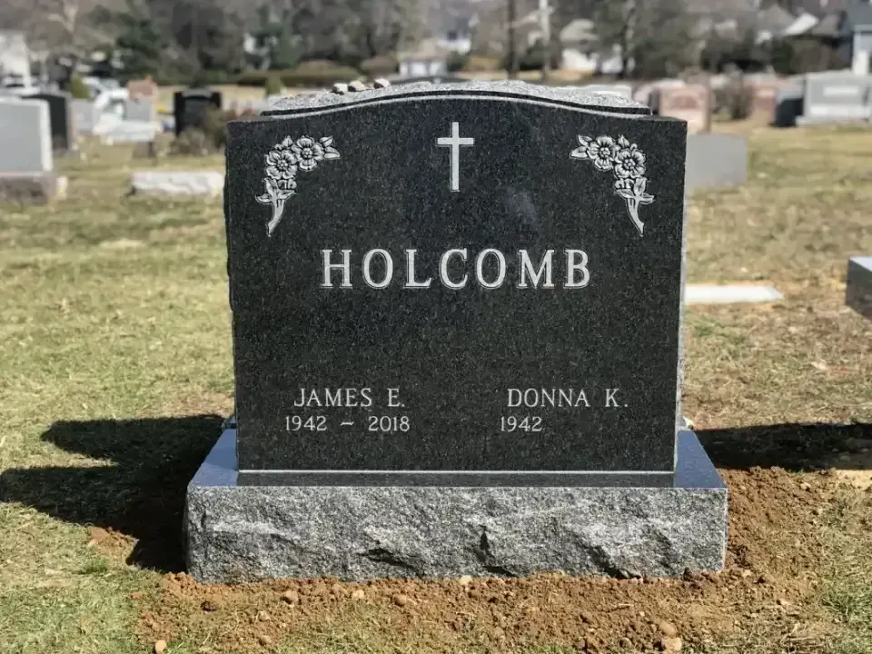 Black granite Holcomb gravestone with cross and floral engraving in a cemetery.