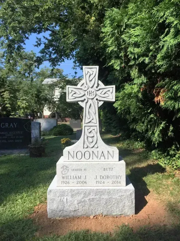 White gravestone with Celtic cross and “NOONAN” in a sunny cemetery, framed by trees.