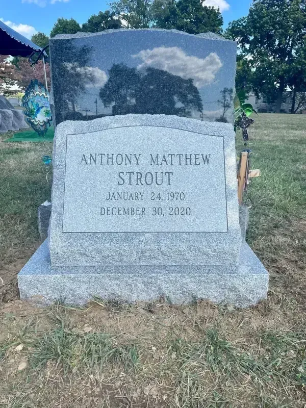 Gray granite gravestone in a grassy cemetery under a cloudy sky, with engraved name and flowers in the background