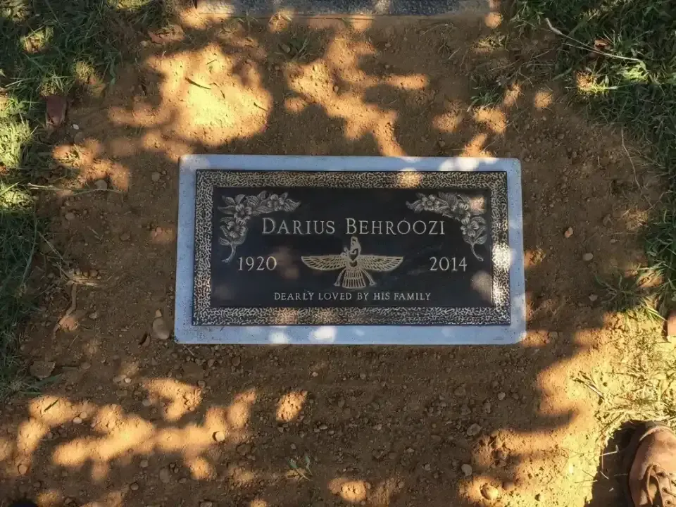 Gravestone plaque in a sun-dappled dirt area, framed in stone, with engraved name and dates.