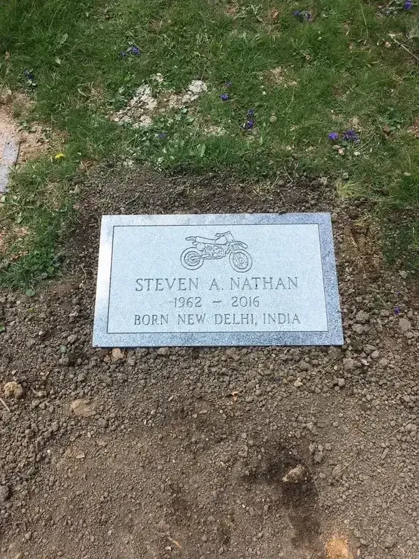Gravestone plaque set in dirt beside grass and small purple flowers