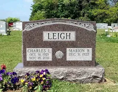 Gravestone reading “LEIGH” in a cemetery with flowers in front and trees in the background