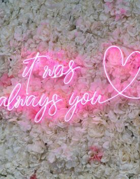 A neon sign that says `` it was always you '' is on a wall of flowers.