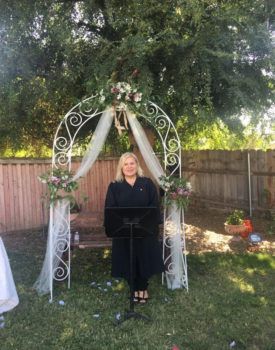 A woman is standing in front of a wedding arch in a backyard.