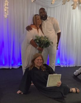 A bride and groom are posing for a picture with a woman laying on the floor holding a certificate.