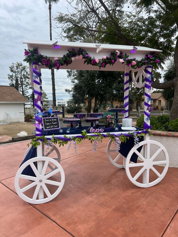 A candy cart decorated in purple and white is sitting on a sidewalk.
