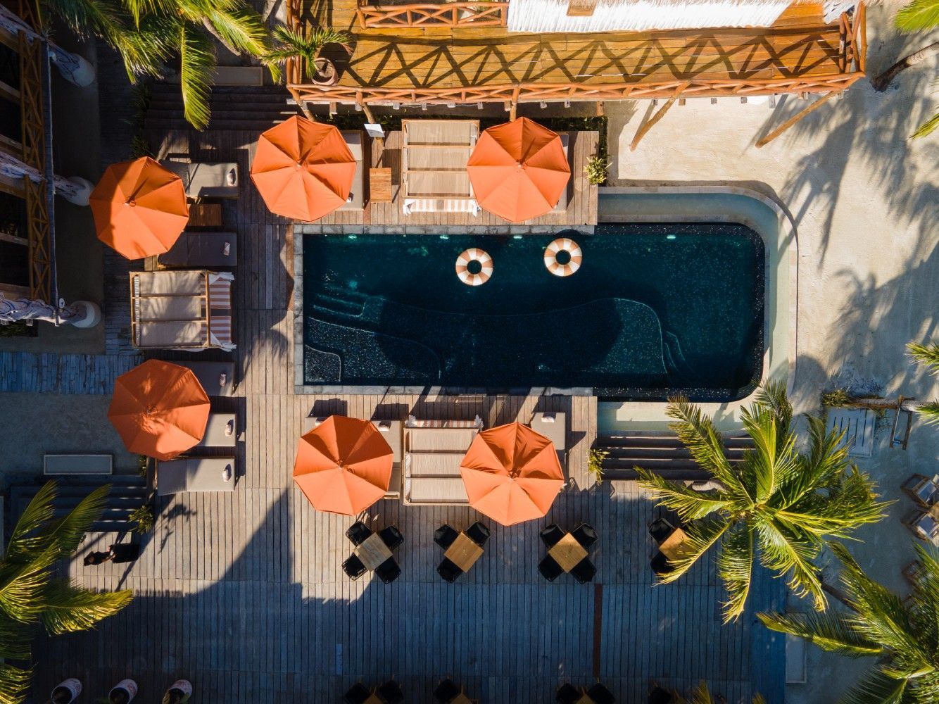 Overhead view of pool area with orange umbrellas, lounge chairs, and tables; palm trees surround.
