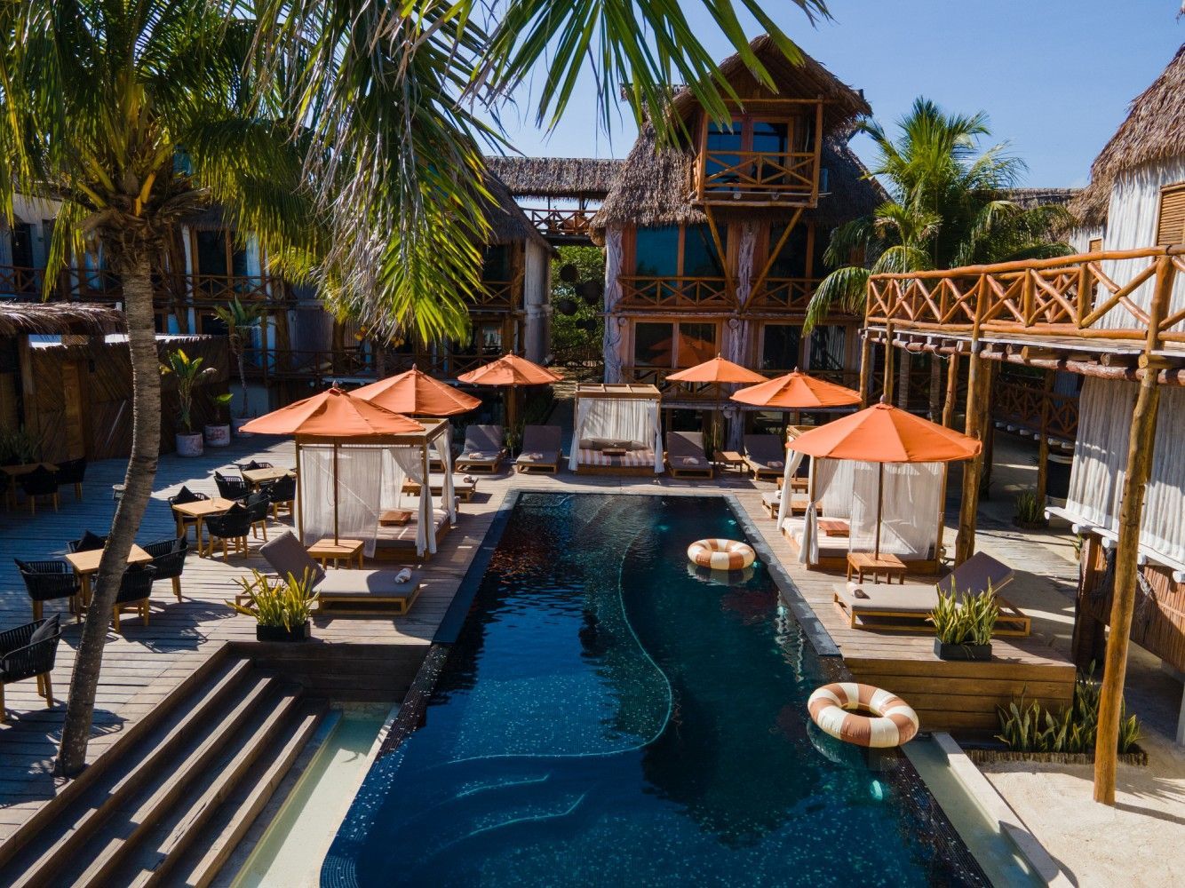Pool with orange umbrellas and cabanas surrounded by tropical buildings and palm trees.