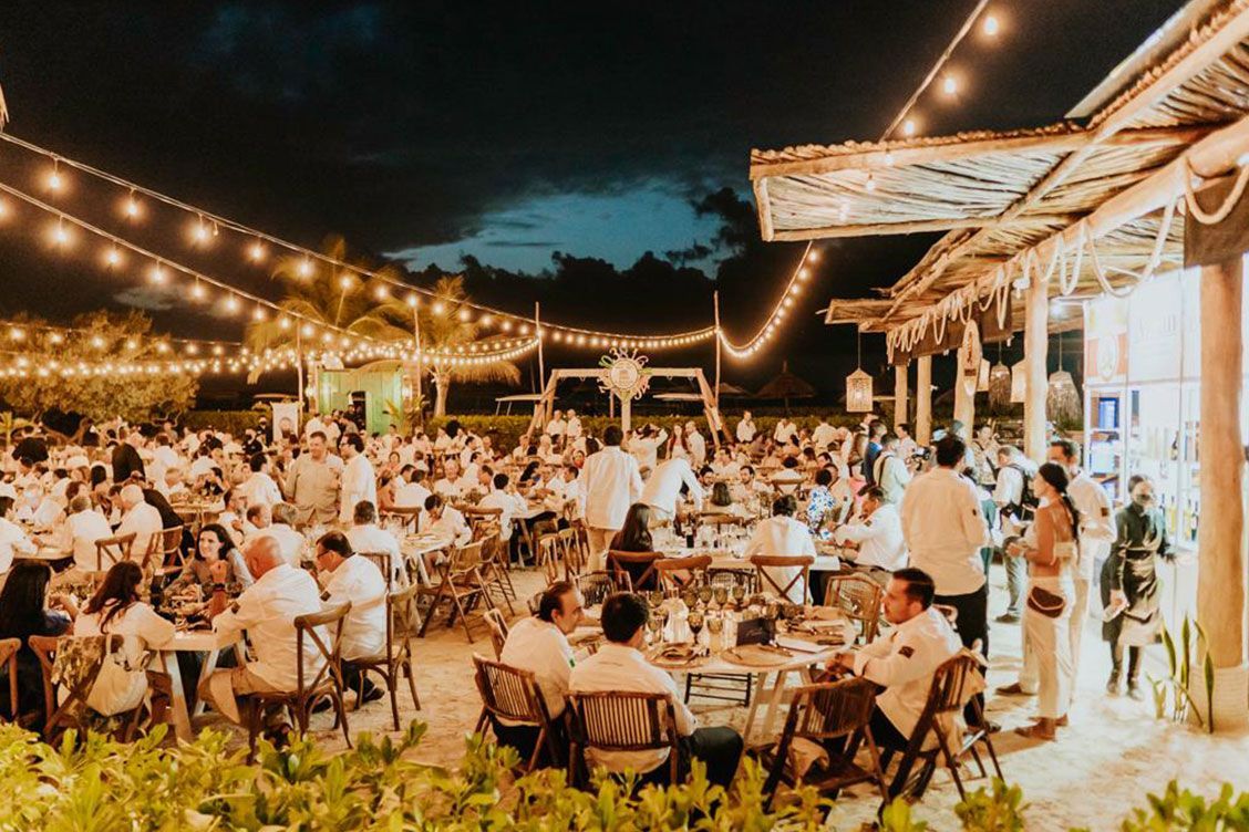 Nighttime outdoor party with many people in white clothing, strung lights, and tables.