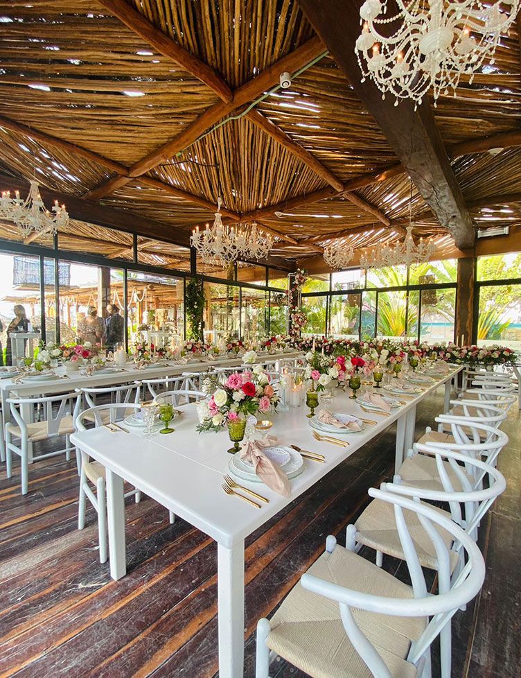 Long white tables set for a celebration with pink floral arrangements, under a wooden roof.